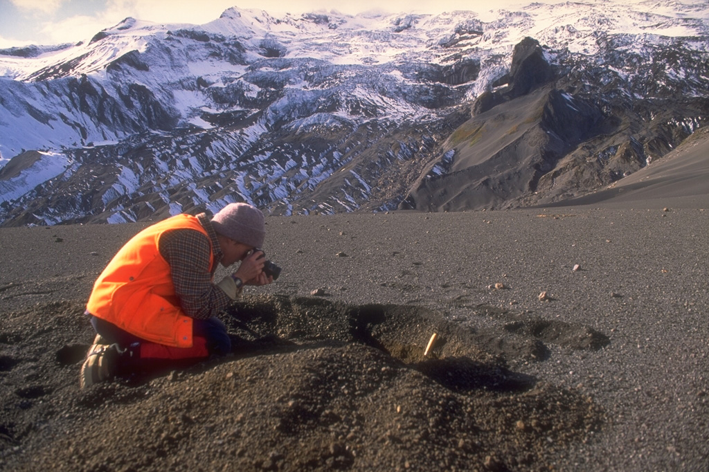 A scientist from the Alaska Volcano Observatory studies tephra from the 1992 eruptions of the Crater Peak vent of Mount Spurr volcano. Three brief explosive eruptions blanketed narrow swaths of the surrounding area with ash. Detailed investigations of deposits are necessary to understand eruption characteristics and magnitude. In this view, about 15 cm of coarse ash and lapilli is exposed in the pit and blocks from the 16-17 September 1992 eruption are scattered across the surface. Photo by Game McGimsey, 1992 (Alaska Volcano Observatory, U.S. Geological Survey).
