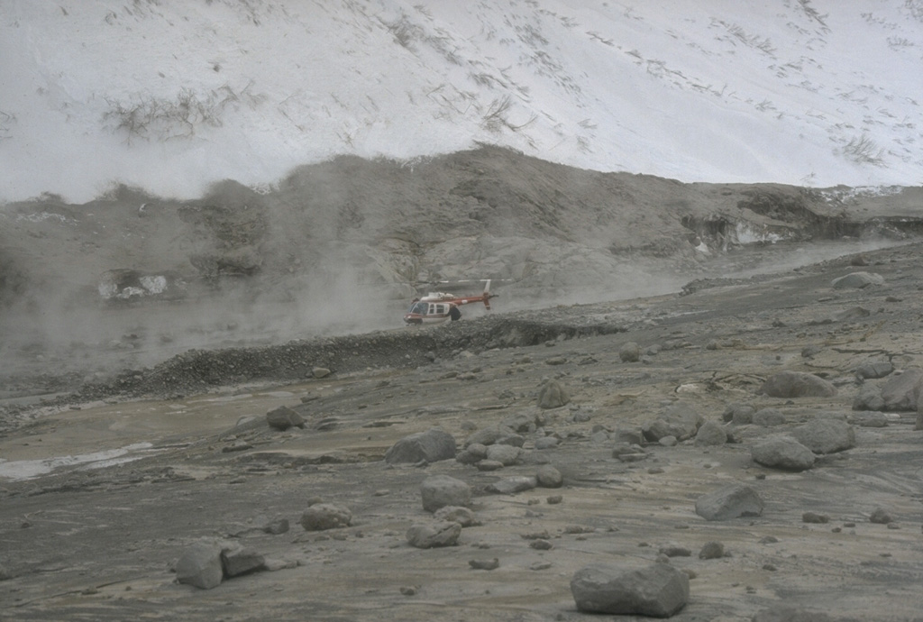 Hot avalanches down the steep north flank of Redoubt during the 1989-90 eruptions mixed with water derived from melted snow and ice to form lahars. These sediment-rich floods carried steaming debris many kilometers down the Drift River Valley. The top of the gray area marks the high-water line of the lahar that contrasts with the snow-covered slopes above it. Note the helicopter for scale. Photo by Tom Miller, 1990 (Alaska Volcano Observatory, U.S. Geological Survey).