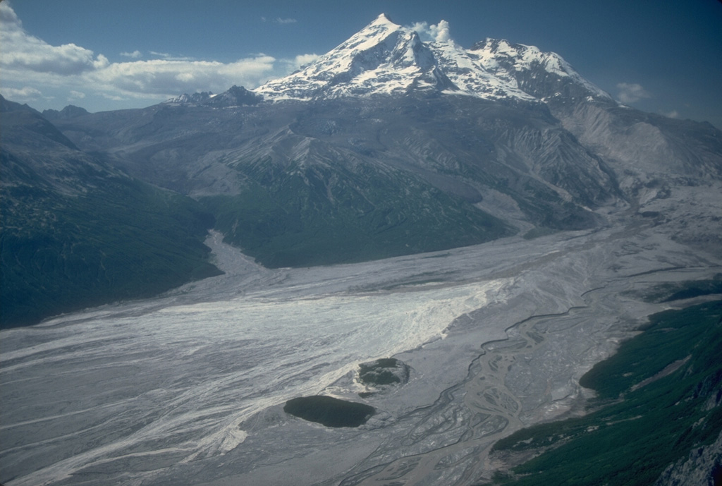 An aerial view shows the upper Drift River valley following the 1989-90 eruptions of Redoubt. Two bedrock islands (informally called the "Dumbbell Hills") are visible at the bottom-center, surrounded by fresh lahar deposits. This photo was taken from the NE on 28 June 1990, shortly after the end of the eruption. Photo by Cynthia Gardner, 1990 (U.S. Geological Survey).