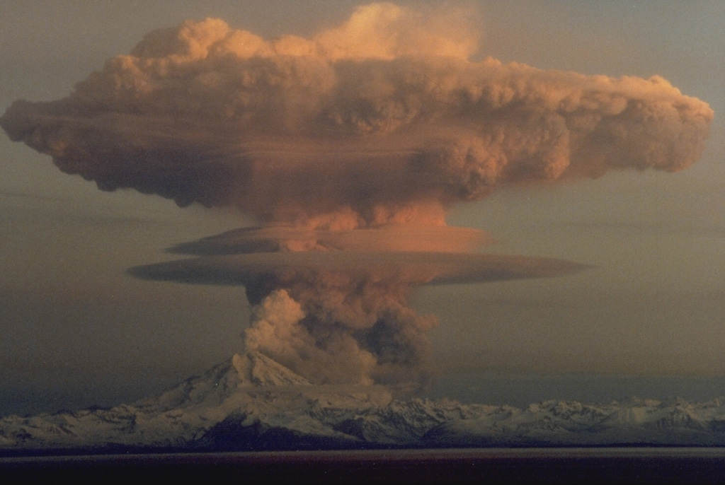 A series of powerful eruptions at Redoubt began on 14 December 1989. This 18 December view from the Kenai Peninsula across Cook Inlet shows an umbrella cloud that appears to originate from a vent on the N flank, but is actually ash from a pyroclastic flow that is traveling down the Drift River valley to the north. Ash plumes during the initial days of the eruption reached heights of about 10 km. Several episodes of strong explosive activity and lava dome growth lasted until June. Copyrighted photo by Robert Clucas, 1990.