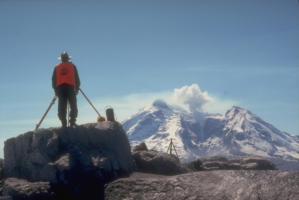 An Alaska Volcano Observatory geologist uses a laser-surveying instrument to measure precise distances to targets installed on the flanks of Redoubt. Minute changes in distances to the targets can reflect ground deformation that may indicate magma movement or other processes. Steam rises above a lava dome in the crater of Redoubt in this photo taken on 5 May 1990, near the end of an eruption that had begun the previous December. Photo by Game McGimsey, 1990 (Alaska Volcano Observatory, U.S. Geological Survey).