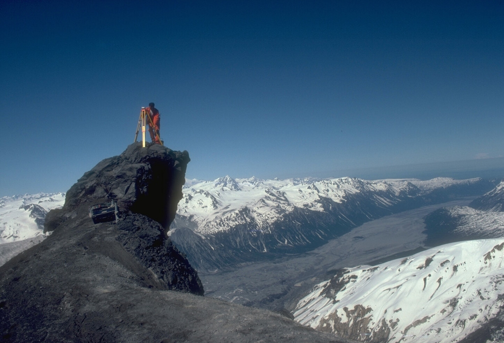 An Alaska Volcano Observatory geologist sets up GPS (Global Positioning System) instrumentation on the N flank of Redoubt volcano. The GPS receiver calculates an extremely accurate location through satellite-based triangulation. This helps pinpoint locations for electronic distance measurements that detect deformation that may be related to eruptive activity. The Drift River valley extending away from the volcano to the NE was covered with pyroclastic-flow and mudflow deposits from the 1989-90 eruption. Photo by Game McGimsey, 1991 (Alaska Volcano Observatory, U.S. Geological Survey).