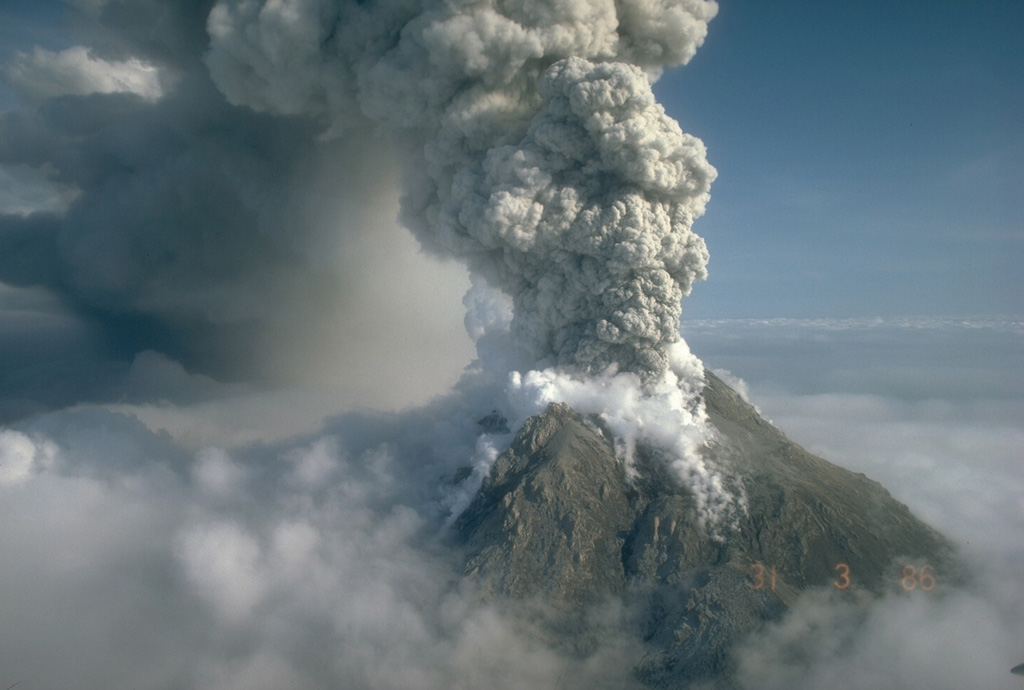 An ash plume rises above Augustine on 31 March 1986, eventually reaching a height of 12 km. Powerful explosions during the initial days of the 1986 eruption, which began on 27 March, removed portions of the 1976 summit lava dome. This view from the SW shows a darkened area of ash deposition downwind from the eruption plume. Photo by Betsy Yount, 1986 (Alaska Volcano Observatory, U.S. Geological Survey).