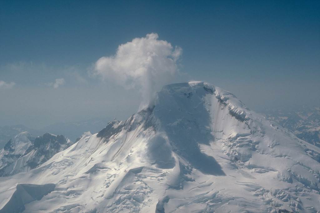 The north face of Iliamna, one of the Cook Inlet volcanoes monitored by the Alaska Volcano Observatory, is seen in this 6 May 1986 aerial view. Steam and volcanic gases rise from the near-continuously active fumaroles high on the eastern and southern flanks. Photo by Game McGimsey, 1986 (U.S. Geological Survey, Alaska Volcano Observatory).