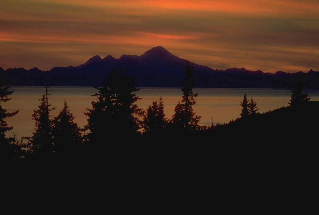 Iliamna rises across Cook Inlet, seen here at sunset from the Kenai Peninsula. It is the southernmost of three large volcanoes west of the inlet. The eroded summit ridge consists of four peaks along a 5-km-long ridge extending south (left) of the highest point. Photo by Alaska Volcano Observatory, U.S. Geological Survey.