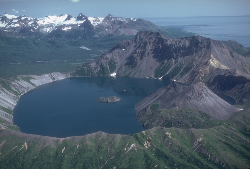 The small 2.5-km-wide Kaguyak caldera is filled by a lake that reaches 550 m below the caldera rim, seen here from the west. A lava dome extends into the lake on the SW side and another dome forms a small island in the center of the lake. The voluminous caldera-forming deposits have been radiocarbon dated at 5,800 years old. A large pre-caldera lava dome forms the high point on the eastern caldera rim. The broad valley of Big River descends to Shelikof Strait to the upper right. Photo by Chris Nye, 1982 (Alaska Division of Geological and Geophysical Surveys, Alaska Volcano Observatory).