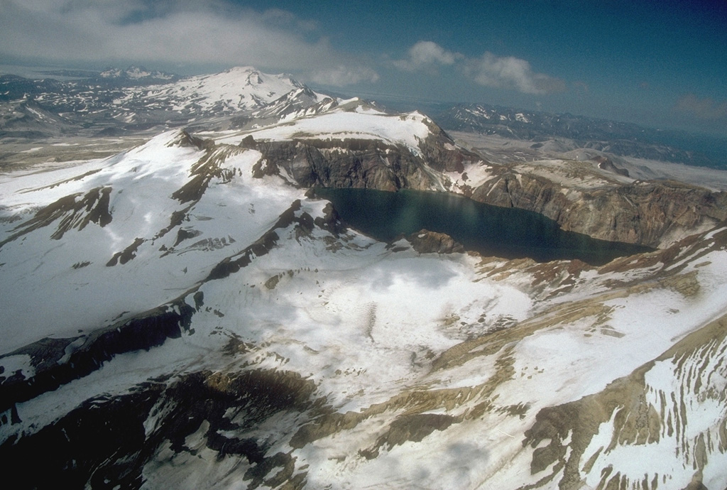 Katmai caldera, formed during the catastrophic eruption of the nearby Novarupta vent in June 1912, is seen here in an aerial view from the NE. The steep-walled, 1.5-km-wide caldera formed on a cluster of overlapping cones and is partially filled by a lake. Beyond the caldera are the multiple peaks of Trident volcano; Mageik volcano is the snow-and-ice-covered cone in the background. Photo by Chris Nye, 1991 (Alaska Division of Geological and Geophysical Surveys, Alaska Volcano Observatory).