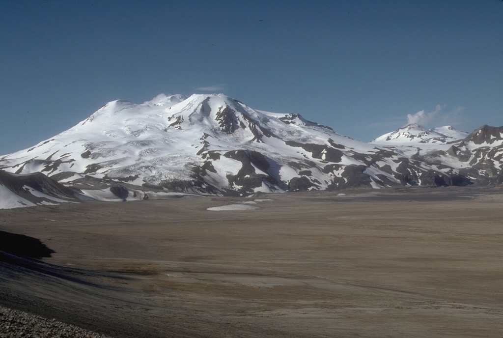 Mount Mageik (left) and Mount Martin (distant right) tower above the flat floor of the Valley of Ten Thousand Smokes. The deposits in the valley are pyroclastic flows that formed during the 1912 eruption of Novarupta, the largest eruption of the 20th century. Glacier-covered Mageik has a broad summit containing multiple cones and vents. Photo by Game McGimsey (U.S. Geological Survey, Alaska Volcano Observatory).
