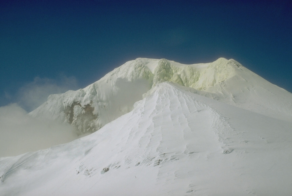 Mount Martin is located at the southern end of the group of volcanoes in the Katmai area. Gases rise from the summit crater and sulfur has accumulated on the snow and ice in this 1990 view. The crater, which opens to the east, is the site of intense fumarolic activity and sometimes contains a small crater lake. Photo by Christina Neal, 1990 (U.S. Geological Survey, Alaska Volcano Observatory).