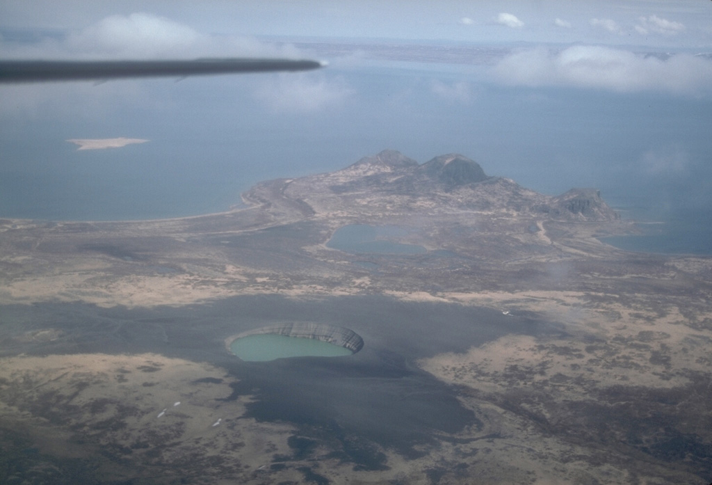 Ukinrek Maars are two phreatomagmatic explosion craters that were created in an area without previous volcanic activity during a 10-day-long eruption that began on 30 March 1977. A greenish lake fills East Maar, the largest of the two in this 1994 view from the south. The smaller West Maar is surrounded by dark tephra near the left margin. Gas Rocks, an older volcanic center, forms the peninsula extending into Becharof Lake in the background. Photo by Chris Nye, 1994 (Alaska Division of Geological and Geophysical Surveys, Alaska Volcano Observatory).