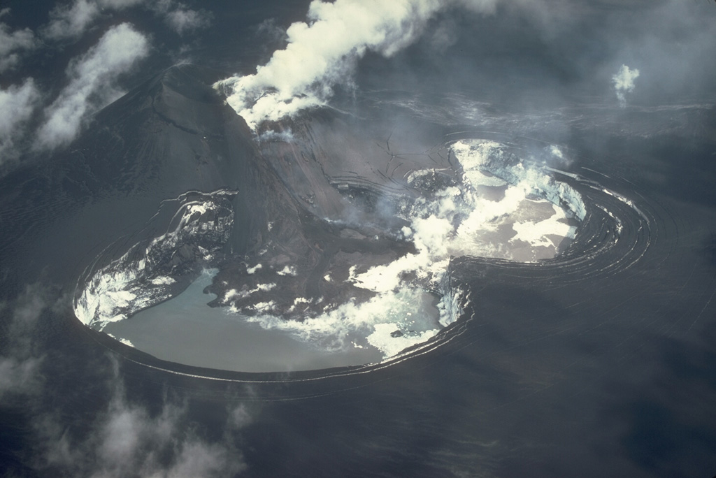 Strombolian eruptions from a scoria cone (upper left) produced heavy ashfall that darkens the entire Veniaminof caldera floor in this 26 July 1983 view from the SW. A plume rises above a vent near the top of the cone that is producing a lava flow down the flank and melting a large pit in the glacial ice Two meltwater lakes have formed in the depressions and steam rises from the margins of the lava flow. Concentric fractures are visible around the margin of the pit due to exposed glacial ice from the collapse. Photo by Betsy Yount, 1983 (Alaska Volcano Observatory, U.S. Geological Survey).