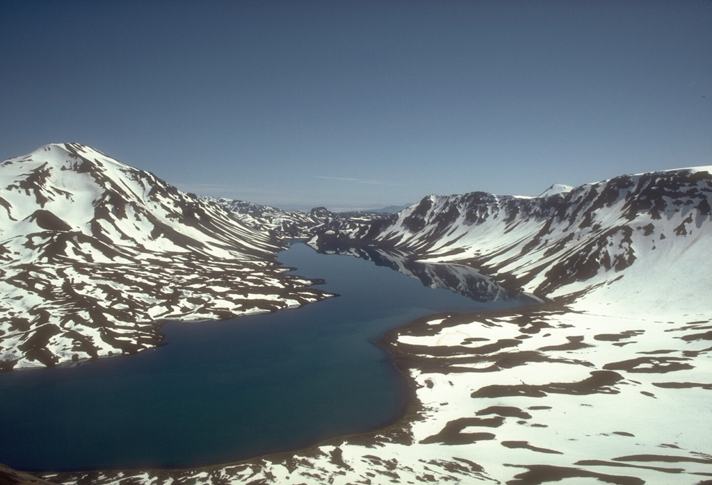 Emmons Lake is a 11 x 18 km caldera and is one of the largest calderas in the Aleutian arc. The caldera formed during two large Quaternary eruptions that produced welded tuffs extending to both the Bering Sea and Pacific Ocean. This 1988 view from the SW shows Emmons Lake, the southern caldera wall (right), and Mount Emmons, a post-caldera volcano (left). The post-caldera  Mount Emmons, Double Crater, and Mount Hague cones are oriented along the same NE trend as the elongate caldera. Photo by Tom Miller, 1988 (Alaska Volcano Observatory, U.S. Geological Survey).