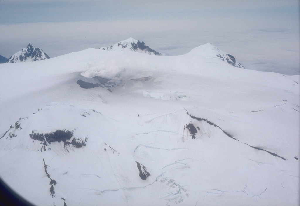 An aerial view of the summit area of Makushin volcano in the northern part of Unalaska Island in the eastern Aleutians, shows a small plume rising from a crater (left center). The broad summit contains a 2.5-km-wide Holocene caldera with a scoria cone that has produced minor explosive eruptions in historical time. Photo by Chris Nye, 1982 (Alaska Division of Geological & Geophysical Surveys).
