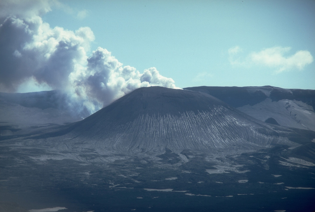 A steam-and-gas plume rises in December 1980 from a scoria cone in Alaska's Okmok caldera. Known only as Cone A, it is one of numerous unnamed cones within the 10-km-wide caldera. Erosional furrows produce linear stripes on the flank of the cone. Eruptions in 1945 and 1958 produced lava flows onto the caldera floor. The wall of Okmok caldera forms the skyline ridge. Photo by Chris Nye, 1980 (Alaska Division of Geological & Geophysical Surveys, Alaska Volcano Observatory).
