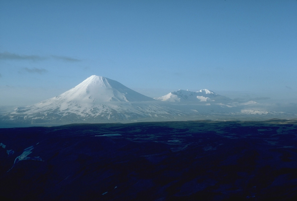 The effects of erosion are visible in this view from the SW of two prominent stratovolcanoes on SW Umnak Island in the Aleutians. Mount Vsevidof (left) is a near-symmetrical volcano where frequent eruptions, which have continued into historical time, have overcome the effects of erosion. In contrast, Recheschnoi volcano (right) has been inactive for longer periods of time and has been extensively dissected by glaciers. Only small pyroclastic cones and lava domes have erupted during the past 10,000 years. Photo by Chris Nye, 1985 (Alaska Division of Geological & Geophysical Surveys).