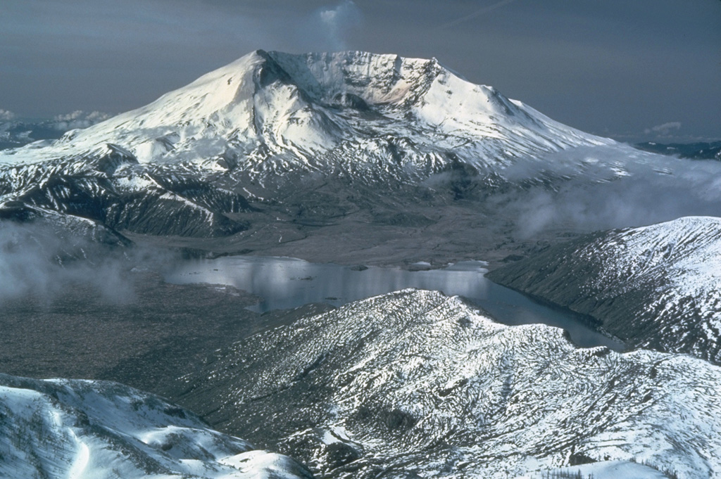 The 1980 eruption of Mount St. Helens dramatically altered the conical profile of the volcano. The eruption left a 2 x 3.5 km wide horseshoe-shaped crater, seen here from the NNE beyond Spirit Lake (center). The landslide of 18 May lowered the summit by 400 m and produced a highly mobile debris avalanche that swept into Spirit Lake and traveled down the North Fork Toutle River. The associated northward-directed lateral blast devastated about 600 km2. Photo by Lyn Topinka, 1981 (U.S. Geological Survey, Cascades Volcano Observatory).