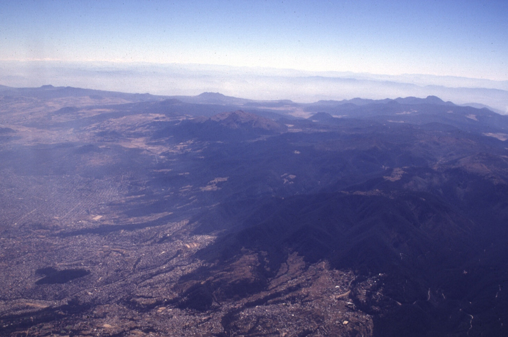 Mexico City encroaches onto the Chichinautzin volcanic field, which covers a broad 1,000 km2 area immediately south of the city. Many of the more than 150 small cones in the volcanic field are of Holocene age. Among the larger features of the field seen in this aerial view from the NW are Volcán Ajusco (the brown-colored peak in the center below the skyline) along with Cerro Pelado, Cerro Chichinautzin, and Cerro Tláloc to the left. In addition to the Xitle eruption less than 2,000 years ago, an eruption of Cerro Chichinautzin was witnessed by local inhabitants. Photo by Lee Siebert, 1997 (Smithsonian Institution).