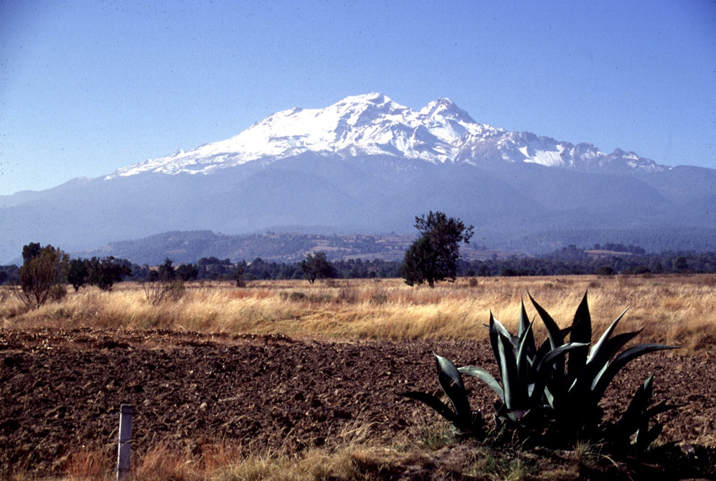 Iztaccíhuatl rises SW of the Puebla basin in central México. A series of overlapping cones constructed along a NNW-SSE trend forms the summit ridge, which is 6 km long just above the snowline. Glaciation has extensively eroded the summit and flanks of the dominantly Pleistocene edifice. The Toltec climbed Iztaccíhuatl and left terracotta, carved stone, jade, and obsidian artifacts just below the summit, forming the highest archeological site in North America. Photo by Lee Siebert, 1997 (Smithsonian Institution).