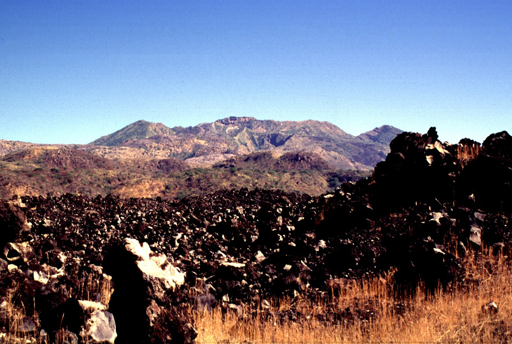 The unvegetated Ceboruco lava flow in the foreground appears to be some of the youngest of the W-flank flows. The peaks on either side of the broad summit in this view from the WSW are the rims of the 4-km-wide outer caldera. Photo by Lee Siebert, 1997 (Smithsonian Institution).