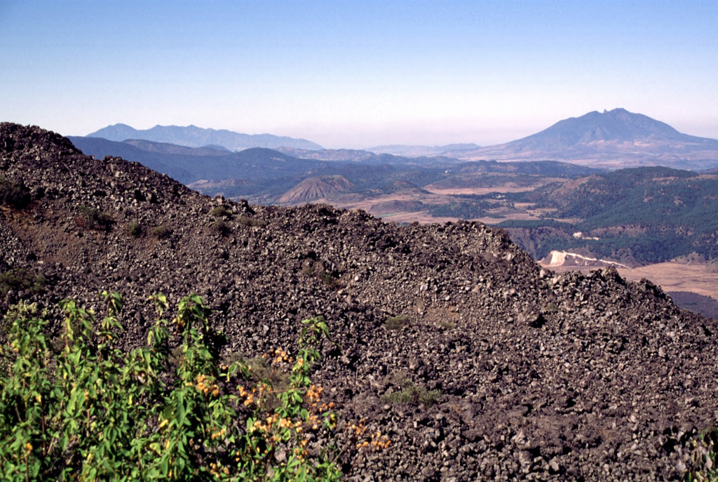 The El Norte lava flows of Ceboruco in the foreground cover much of the northern flank. These young flows erupted from vents within the inner summit caldera and may have been emplaced during one of two 16th-century eruptions. The lava flows traveled 3.5 km down the north flank and formed a broad, 3.5-km-wide front at the base of the volcano. The volcano with the irregular profile in the left distance is Tepetiltic; Sangangüey forms the peak to the far right. Photo by Lee Siebert, 1997 (Smithsonian Institution).