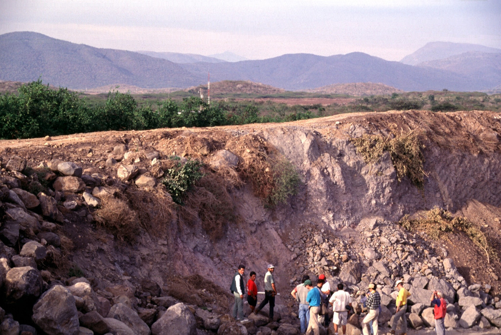 Collapse of México's Colima volcanic complex during the late Pleistocene produced a major debris avalanche and debris flow that traveled 120 km to the Pacific Ocean. The quarried hummock in the foreground and the two brown hills in the middle distance are composed of material transported more than 100 km from Colima volcano (seen in the far distance above the left-hand hummock) and incorporated from valley floors during transport. Photo by Lee Siebert, 1997 (Smithsonian Institution).