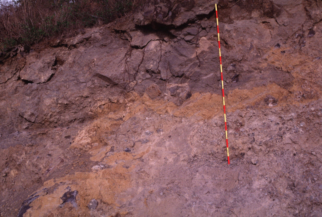 Textures associated with volcanic landslides are preserved in this quarry wall more than 100 km from the source of this major avalanche from México's Colima volcano. Large fractured blocks appear at the top of the exposure, and a color mottling is produced by adjacent segments of the volcano or valley floor material incorporated during transport. A 2-m-high ruler provides scale. Photo by Lee Siebert, 1997 (Smithsonian Institution).