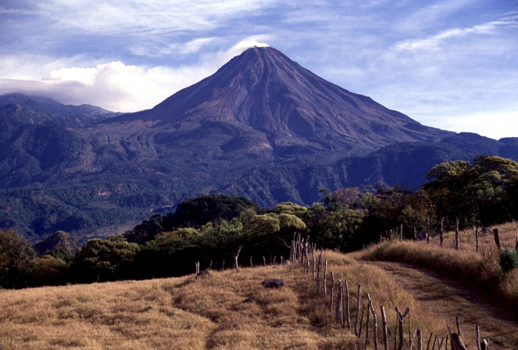 Volcán de Colima was constructed within a large horseshoe-shaped scarp formed by repetitive collapse of a previous edifice. The northern scarp rim forms the arcuate ridge to the far-left of this view from the SW. The modern cone was built over the past 2,600 years and had grown to a height of 400 m above the caldera rim at the time of this 1997 photo. Lava flows form much of the flanks. Photo by Lee Siebert, 1997 (Smithsonian Institution).