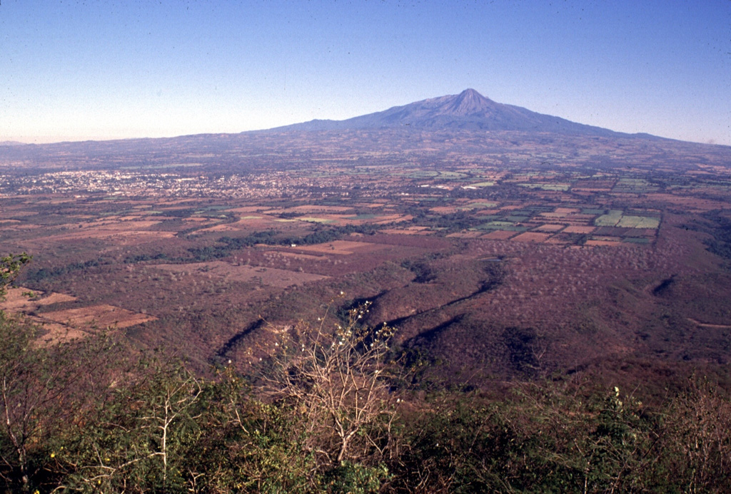 The history of the Colima volcanic complex has been characterized by repetitive collapse of the cones, producing rapid debris avalanches that can travel far from their source. The entire broad apron south of Colima in this view, including the area underlying Colima City to the left, is formed of deposits produced by volcanic landslides and associated lahars. The youngest avalanche, about 2,600 years ago, traveled 35 km as far as the city of Colima.  Photo by Lee Siebert, 1997 (Smithsonian Institution).