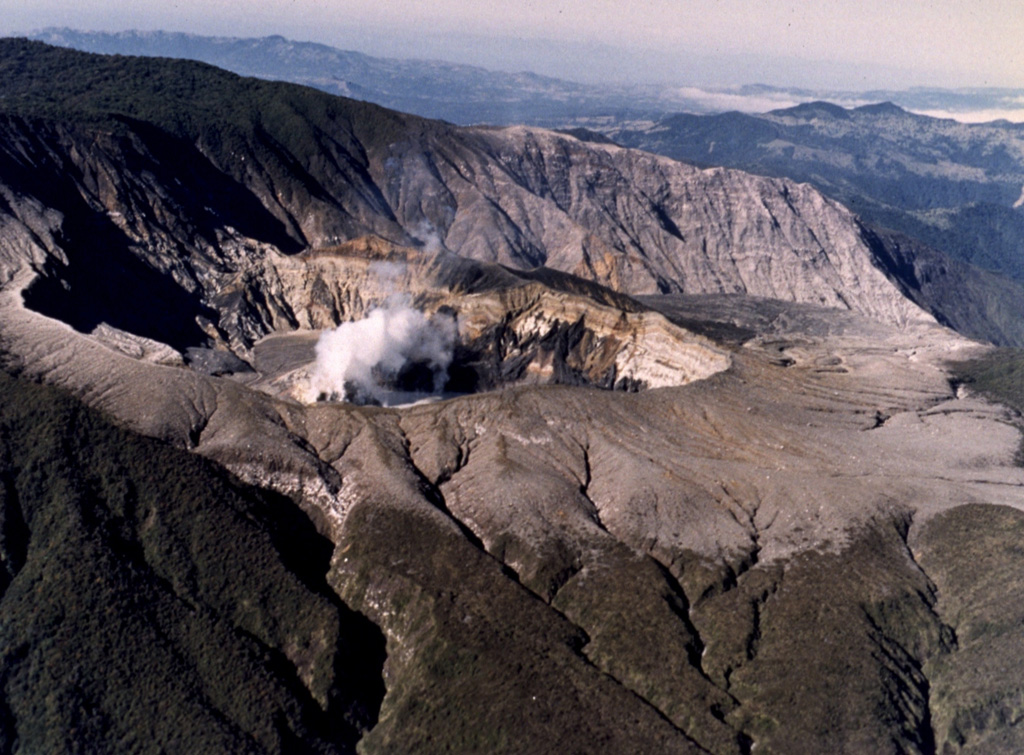 The degassing Poás crater, seen here in an aerial view from the NE, lies within the smaller of two nested calderas at the summit. This inner caldera is 4 km in diameter. A tourist overlook along the ESE crater rim near the upper left provides a view into the crater. Photo by National Aeronautics and Space Administration (NASA), 1982.