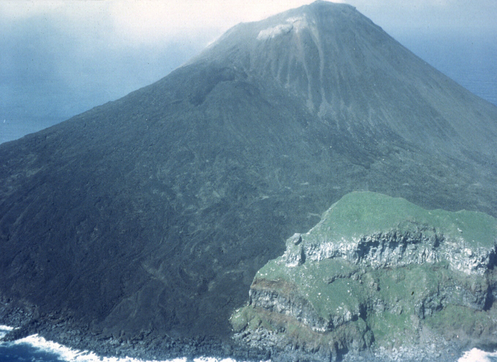 This 1980 photo from the SE of the small island volcano of Farallón de Pájaros shows unvegetated lava flows that diverted around the vegetated peak to the lower right, which is a remant of a caldera that formed prior to the formation of the present cone. Photo by Winfrey, 1980 (U.S. Navy).