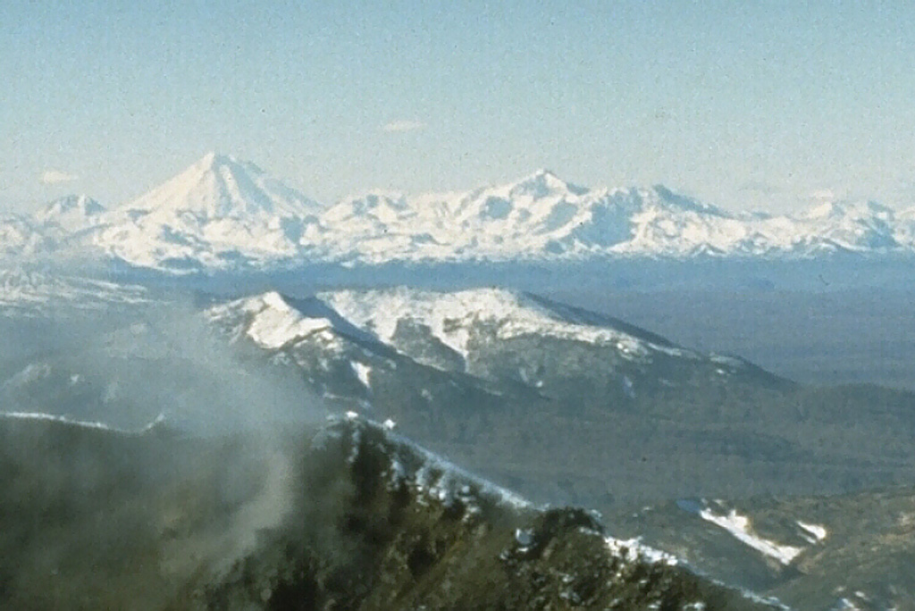 Koryaksky is the tallest peak to the left in the background of this 1990 photo. The ridge to the right of Koryaksky is the eroded Pleistocene age Dzenzursky volcano. This view from the NE has the summit crater rim of Karymsky in the foreground. Photo by Dan Miller, 1990 (U.S. Geological Survey).