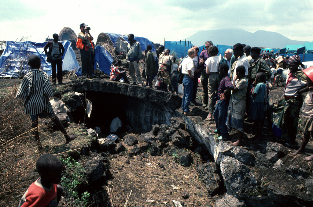 Major humanitarian crises in eastern Africa played out in areas near Nyiragongo and Nyamuragira volcanoes. This August 1994 photo of the Kitumba refugee camp shows a Rwandan family living in a lava tube on a flow on the SW flank of Nyamuragira volcano with tarps of other shelters in the densely occupied refugee camp beyond. Nyiragongo volcano appears on the right skyline. Photo by Jack Lockwood, 1994 (U. S. Geological Survey).
