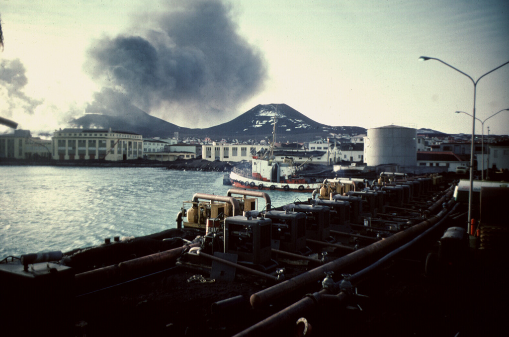 The island of Heimaey, about 10 km off the south coast of Iceland, is part of the Vestmannaeyjar volcanic system, which comprises numerous islands and submarine vents. Heimaey probably formed in the Holocene, through numerous eruptions. Here, an ash-laden eruption plume rises above a volcanic cone behind the town of Vestmannaeyjar in 1973. The water pumping operation in the foreground was designed to cool the front of an advancing lava flow that threatened the town and its harbor. Photo by Jack Lockwood, 1973 (U.S. Geological Survey).