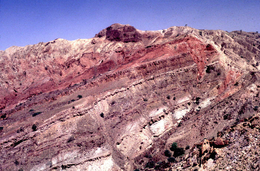 Uplift at Alid has exposed thick sequences of reddish, well-stratified siltstone beds, some very fossiliferous, which accumulated in a tidal or inter-tidal environment. High in the sequence there are some pillow basalts. At the very top of the sequence on the left is a few-meters-thick cover of light-colored rhyolitic Plinian pumice fallout. Photo by Wendell Duffield, 1996 (U.S. Geological Survey).