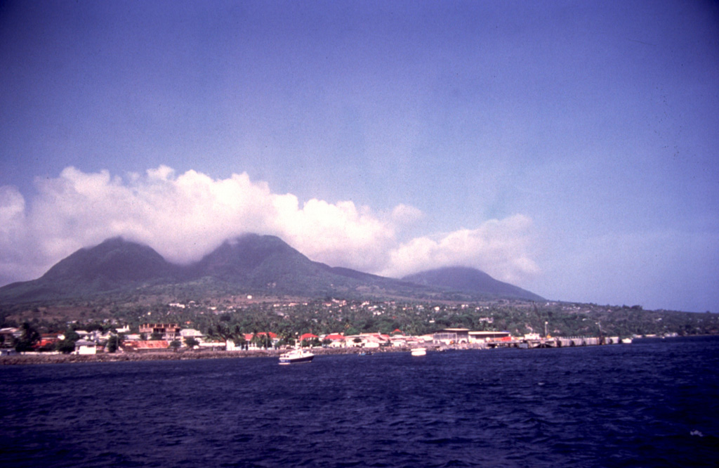 A boat offshore from the former capital city of Plymouth provides a tranquil view of the cloud-draped summit of Soufrière Hills volcano.  The photo was taken in 1994, the year prior to the onset of a major eruption from Soufrière Hills that in 1997 destroyed the city of Plymouth.  The broad low-lying area the city was built on, only 3-4 km from the summit of the volcano, was one of the few flat-lying areas on the small island of Montserrat and had been created by the products of earlier eruptions similar to those that later destroyed the city. Photo by Lydia Pulsipher, 1994 (University of Tennessee).