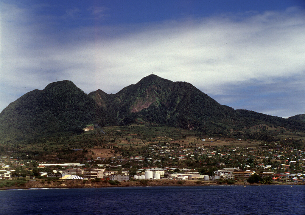Soufrière Hills volcano towers above the peaceful city of Plymouth in 1994, the year prior to the onset of the devastating eruption that eventually destroyed the former capital city.  Pyroclastic flows from a growing lava dome swept through the city to the sea in August 1997 through the low notch (center) in the western crater wall of the volcano.  The city was located only 3 km from the crater rim on top of flat-lying pyroclastic-flow deposits from previous eruptions. Photo by Lydia Mihelic Pulsipher, 1994 (University of Tennessee).