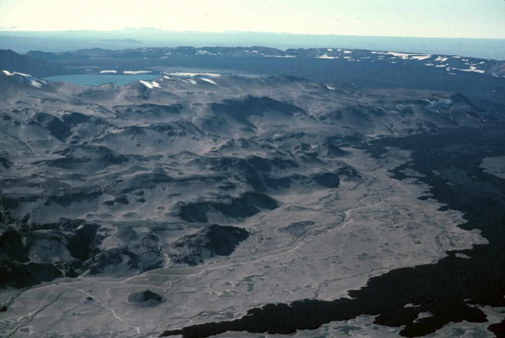 The dark-colored lava flow at the lower right was erupted during 1961 from a vent on the NE side of Askja caldera and flowed about 9.5 km E. The western rim of the ~8-km-diameter main Askja caldera can be seen in the background, rising above its flat, lava-covered floor. The 1961 vents were on the right in this image. The lake at the upper left is Öskjuvatn, which fills Askja's youngest caldera, formed due to the explosive 1875 eruption. The irregular slopes of the Dyngjufjöll massif in the foreground are formed almost entirely by the products of subglacial eruptions. Photo by Michael Ryan, 1984 (U.S. Geological Survey).