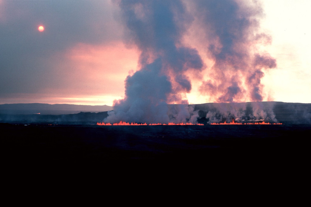 A steam plume rises above a line of small lava fountains on 5 September 1984, the day after the onset of an eruption from an 8.5-km-long fissure extending north from Leirhnjúkur. During the first hours of the eruption, the fissure was active along its entire length, producing a sheet of pahoehoe lava, but later activity was concentrated at a single vent near the northern end of the fissure. This fissure eruption was the last of a decade-long series of Krafla eruptions beginning in 1975 that accompanied an episode of crustal spreading. Photo by Michael Ryan, 1984 (U.S. Geological Survey).