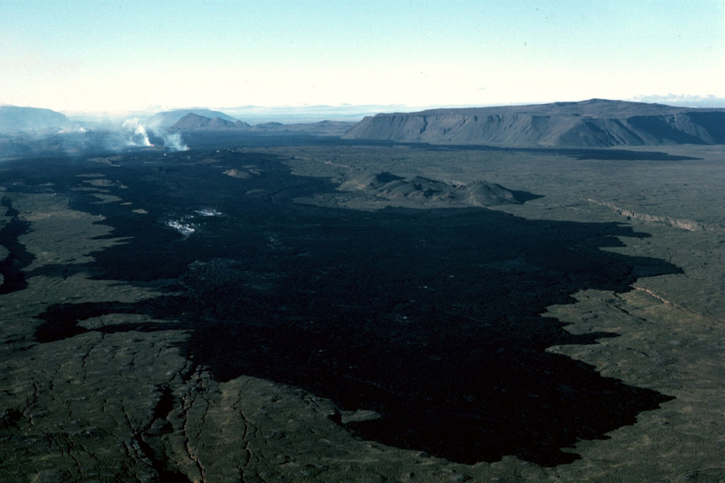 At their northern terminus the 1984 flows form a dark lava field north of Krafla caldera. The northernmost eruptive vents can be seen steaming at the upper left on 10 September 1984, about halfway through the two-week eruption. The flat-topped peak at the upper right is Gæsafjöll, one of Iceland's many table mountains, which formed as a result of repeated eruptions through a glacial icecap. The Hliðarfjall dome can be seen as a peak in the middle-background, and Bláfjall and Sellandafjall table mountains are on the horizon on the left, about 50 km away. Photo by Michael Ryan, 1984 (U.S. Geological Survey).