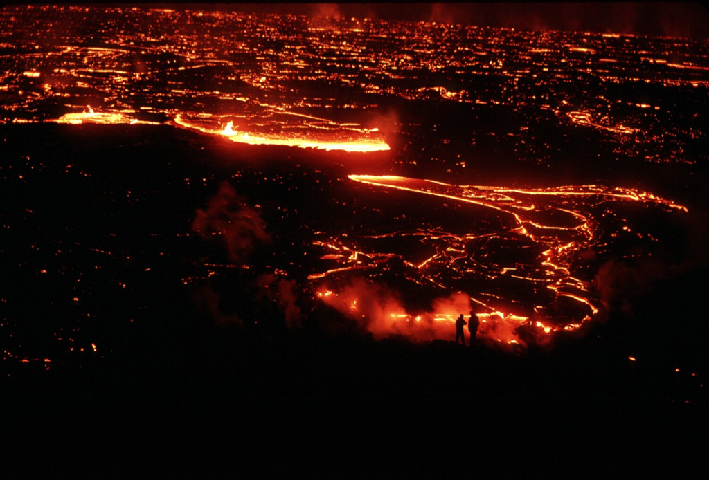 Scientists investigating a lava flow from Krafla volcano are silhouetted (lower right) against the glowing margin of the slowly advancing flow. This photo was taken on 5 September 1984, the day after the onset of an eruption from the Leihrnjúkur fissure. Cracks on the surface of the advancing flow reveal the still-molten interior. Photo by Michael Ryan, 1984 (U.S. Geological Survey).