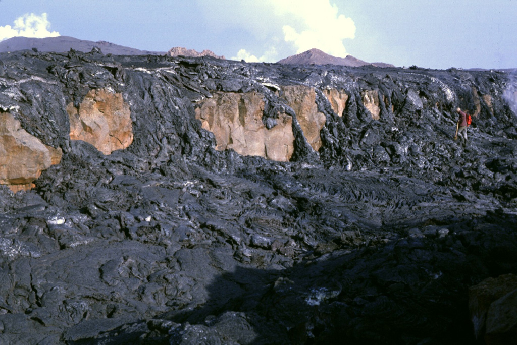 Fresh black pahoehoe lava flows from the 1984 eruption of Krafla drape the Gjastikki fault, whose escarpment forms the brownish cliff surfaces. Note the geologist at the upper right for scale (with red backpack). The still-steaming lava flow, photographed here on 7 September, is only days old and partially buries the fault, which was produced during a previous rifting episode. The steaming cones in the background indicate the location of the vents. Photo by Michael Ryan, 1984 (U.S. Geological Survey).