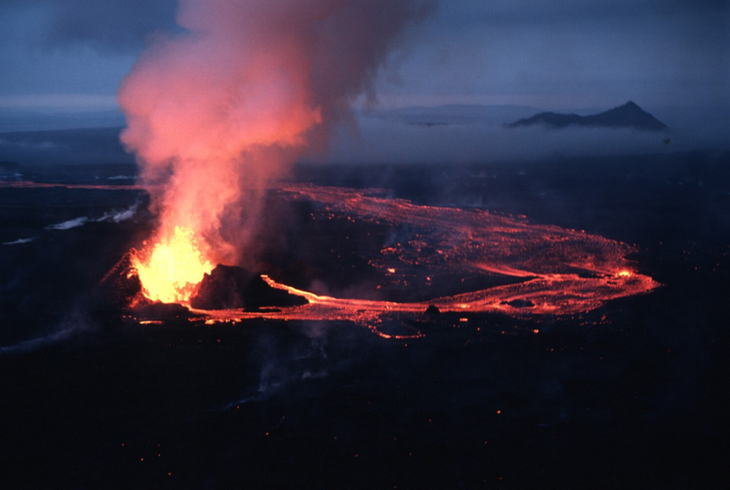 Lava fountains rising from a vent along an eruptive fissure at Krafla feed an incandescent lava flow that travels east and then turns toward the north. This 15 September 1984 photo was taken near the end of a two-week eruption. An 8.5-km-long fissure, initially active along its entire length, fed lava flows that covered 24 km2. The peak rising above the clouds on the right is Eilifur, which likely formed in the Pleistocene. Photo by Michael Ryan, 1984 (U.S. Geological Survey).