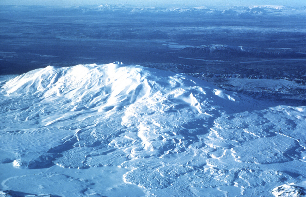 Snow-covered Hekla, one of Iceland's most prominent and active volcanoes, is seen here in an aerial view from the east. Toppgigur crater occupies the summit, in the middle of the ridge, and Axlargigur crater is seen here as a small peak along the summit ridge to the left (W). Repeated eruptions along an ENE-WSW-trending rift are responsible for Hekla's elongated profile. The 5.5-km-long Heklugja fissure cuts across the volcano. Lava flows from eruptions dating back to 1104 CE cover much of the volcano's flanks and are seen here fanning out from the ridge in all directions. Photo by Oddur Sigurdsson, 1977 (Icelandic National Energy Authority).
