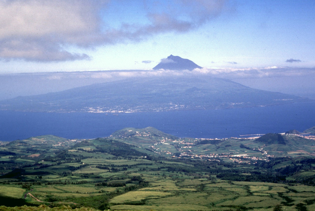The conical Pico stratovolcano rising above the clouds is seen here from Fayal Island to the northwest. The 46-km-long Pico Island can be broadly separated into the Pico stratovolcano in the west, the Planalto da Achada fissure zone in the east (a roughly 30-km-long linear ridge of fissures and cones), and the Topo-Lajes volcano in the south. Pico volcano is capped by a 560-m-wide summit crater that contains a small steep-sided cone visible on the left side of the summit in this image. Recent eruptions have been recorded from the flanks of Pico and in the Planalto da Achada fissure zone.  Photo by Rick Wunderman, 1997 (Smithsonian Institution).