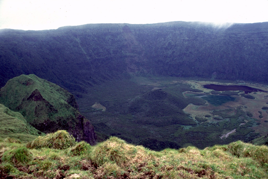 Fayal volcano is capped by a 2-km-wide, 400-m-deep summit caldera, seen here from its southern rim. A small pyroclastic cone (center) and a dark lake (right) can be seen on the floor of the caldera. The caldera was formed incrementally, beginning with a large eruption about 1,000 years ago. Thick airfall-pumice and pyroclastic-flow deposits related to this eruption cover the island. Photo by Rick Wunderman, 1997 (Smithsonian Institution).