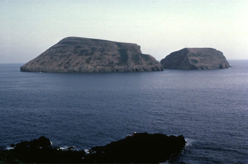 The Ilheus das Cabras islands are the remnants of a tuff cone located 2 km off the southern coast of Terceira Island.  Only the northern half of the tuff cone remains above sea level.  The cone has been eroded into two small islands separated by a narrow channel.  The crater of the tuff cone faces south and is obscured from this view taken west of Ponta da Cruz on the SE coast of Terceira. Copyrighted photo by Katia and Maurice Krafft, 1980.