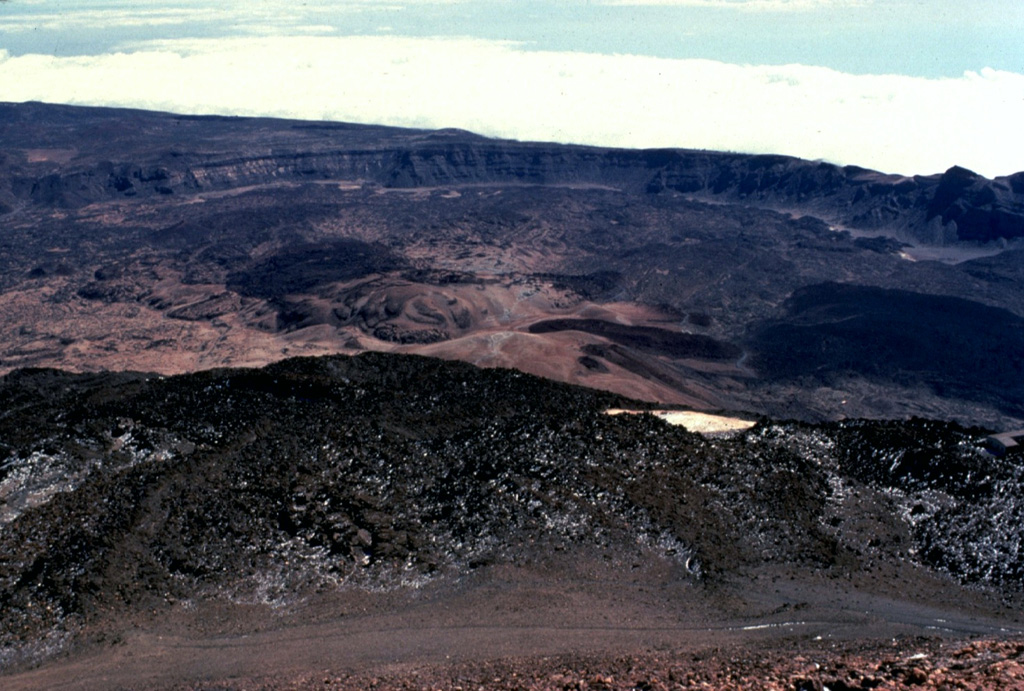 The arcuate rim of Las Cañadas caldera on Tenerife volcano in the Canary Islands rises in the distance above the flat-lying caldera floor.  This view looks to the SE from the summit of Pico de Teide, a large stratovolcano constructed within the massive 10 x 17 km wide caldera.  Dark-colored lava flows from Pico de Teide and other post-caldera cones mantle the caldera floor, which lies 300-m below the far caldera rim and 1700-m below the summit of Pico de Teide.  Copyrighted photo by Katia and Maurice Krafft, 1977.