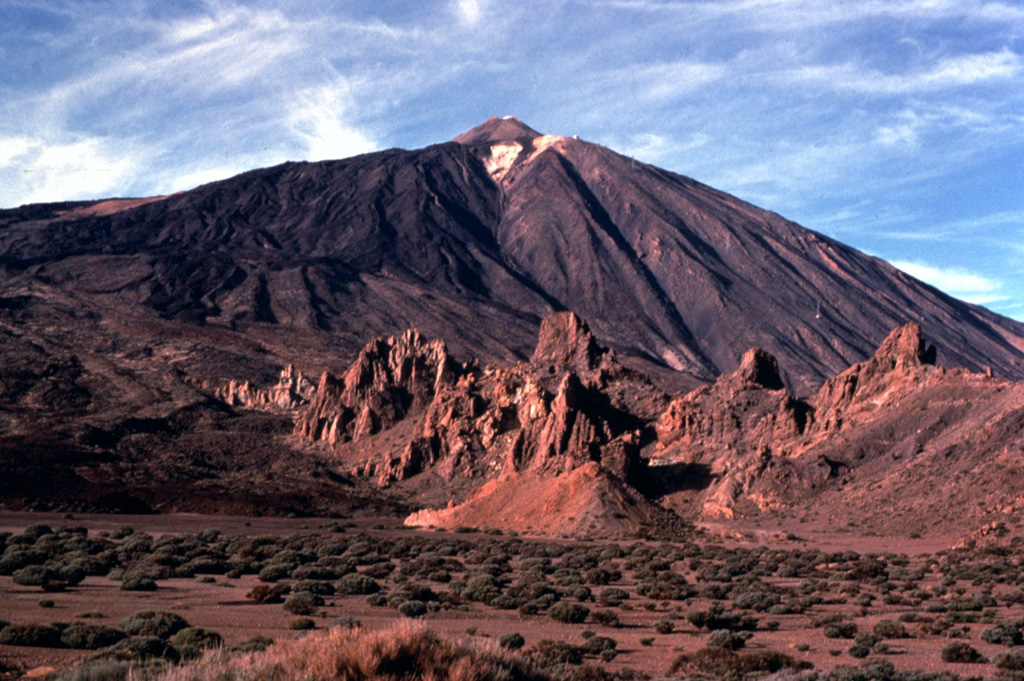 Pico de Teide towers above the Llanos de Ucanca plain on the floor of Las Cañadas caldera.  The rocky spires in the foreground are eroded remains of intrusive phonolitic rocks of the Cañadas formation, predating formation of the caldera.  Youthful dark-colored lava flows blanket the SW (left) side of Teide.  The upper limit of the snowfield just below the summit of Teide marks the rim of the summit crater. Copyrighted photo by Katia and Maurice Krafft, 1977.