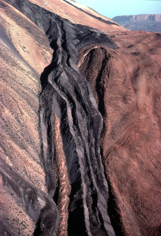 A dark-colored trachytic lava flow descends the flanks of Pico de Teide volcano on Tenerife Island in the Canary archipelago.  The viscous obsidian-bearing lava flow displays steep-sided lateral levees.  These define individual lobes that diverged around a high point on the surface of the flow.  Another lava flow, one of many youthful flows erupted from Pico de Teide, forms the dark streak at the top center of the photo just below the skyline. Copyrighted photo by Katia and Maurice Krafft, 1977.