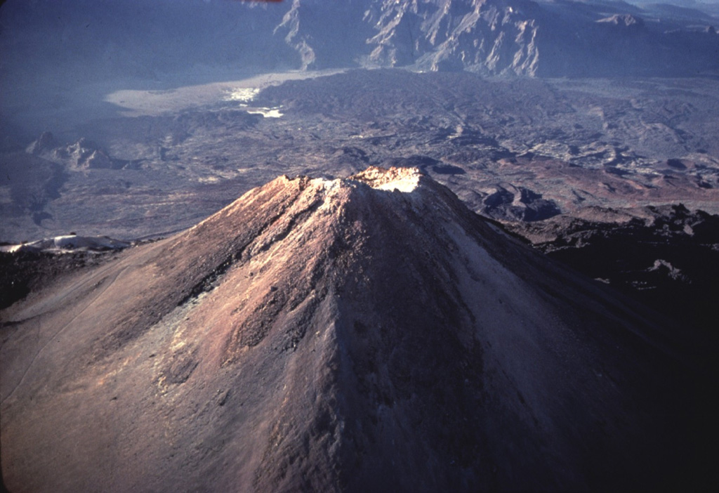 The summit of Pico de Teide volcano towers 1700 m above the floor of Las Cañadas caldera, whose southern caldera wall forms the craggy cliffs at the top of the photo.  Fresh-looking unvegetated lava flows descend 7 km from the summit of Pico de Teide to the caldera wall, spreading across the light-colored sediments (upper left) on the broad caldera floor. Copyrighted photo by Katia and Maurice Krafft, 1977.